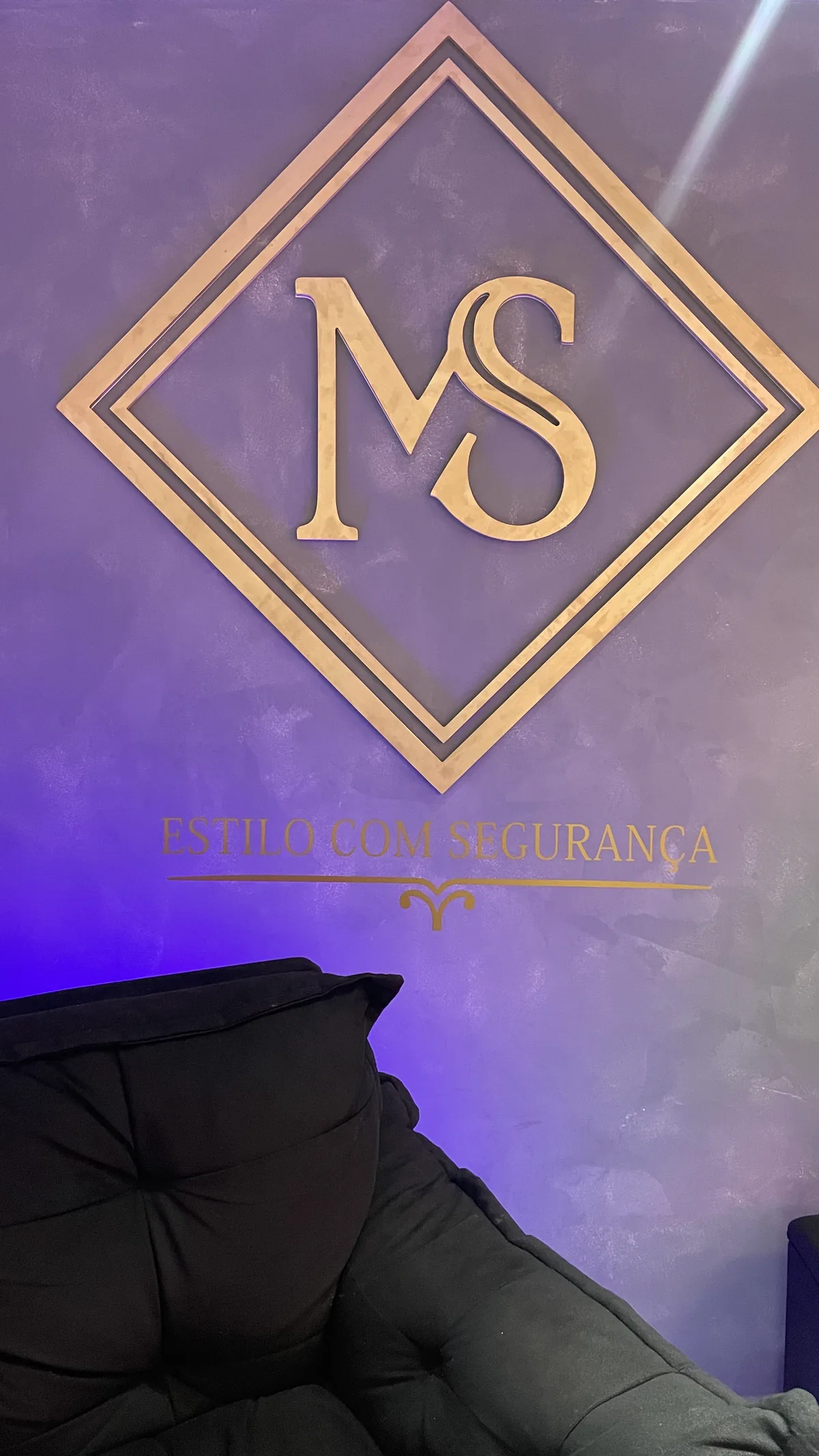 Logo MS em dourado na parede roxa, com a frase Estilo com segurança