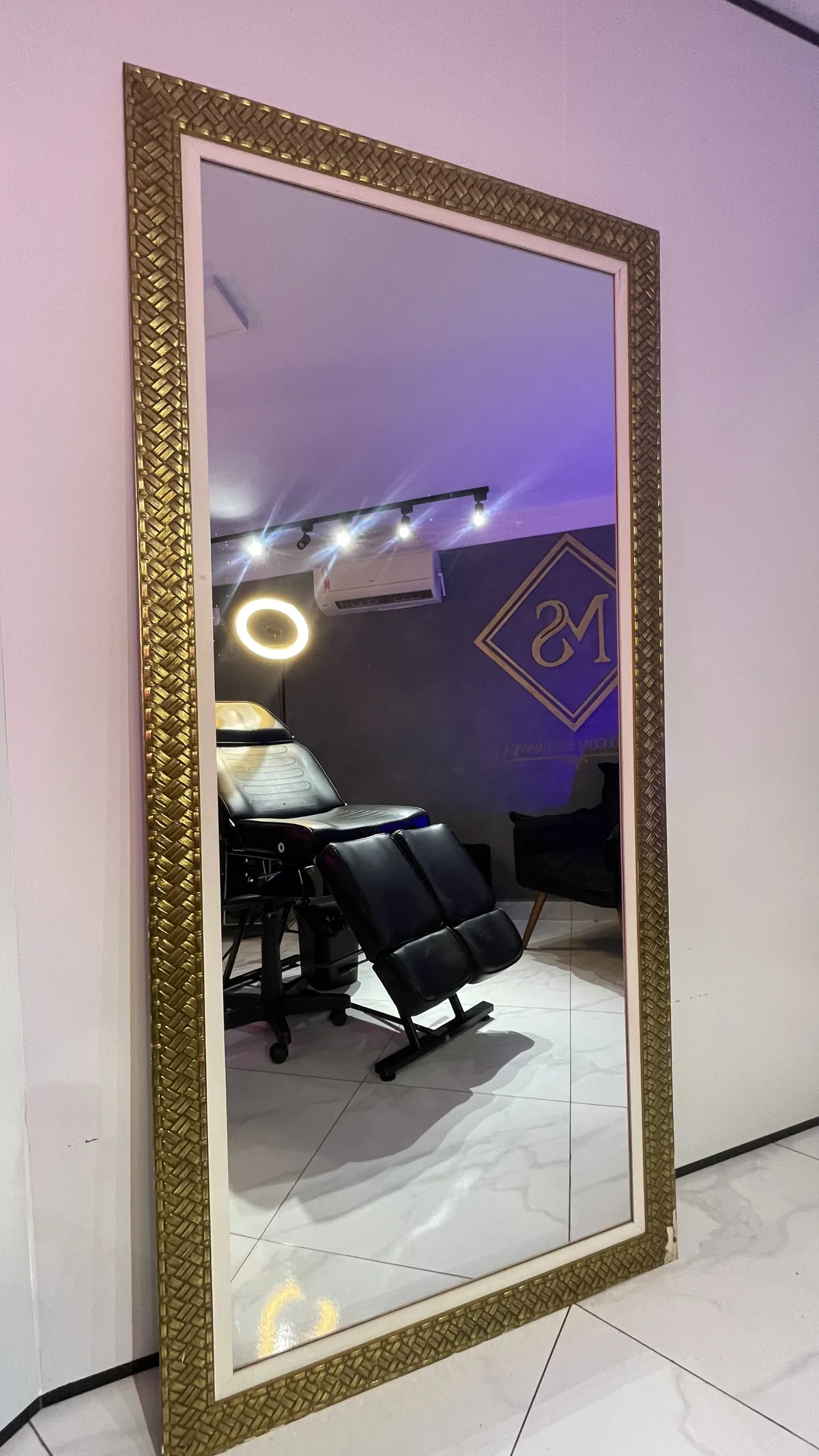 Espelho dourado refletindo a sala de atendimento, a maca e o logo MS na parede